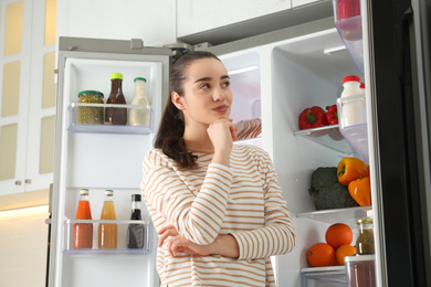 Photo of Thoughtful young woman near open refrigerator indoors