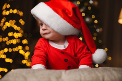 Cute little baby in Santa Claus suit against blurred festive lights. Christmas celebration