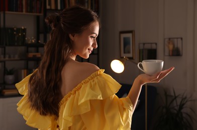 Photo of Beautiful young woman in yellow dress holding cup at home