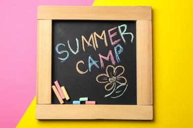 Small blackboard with text SUMMER CAMP, drawing and chalk sticks on color background, top view