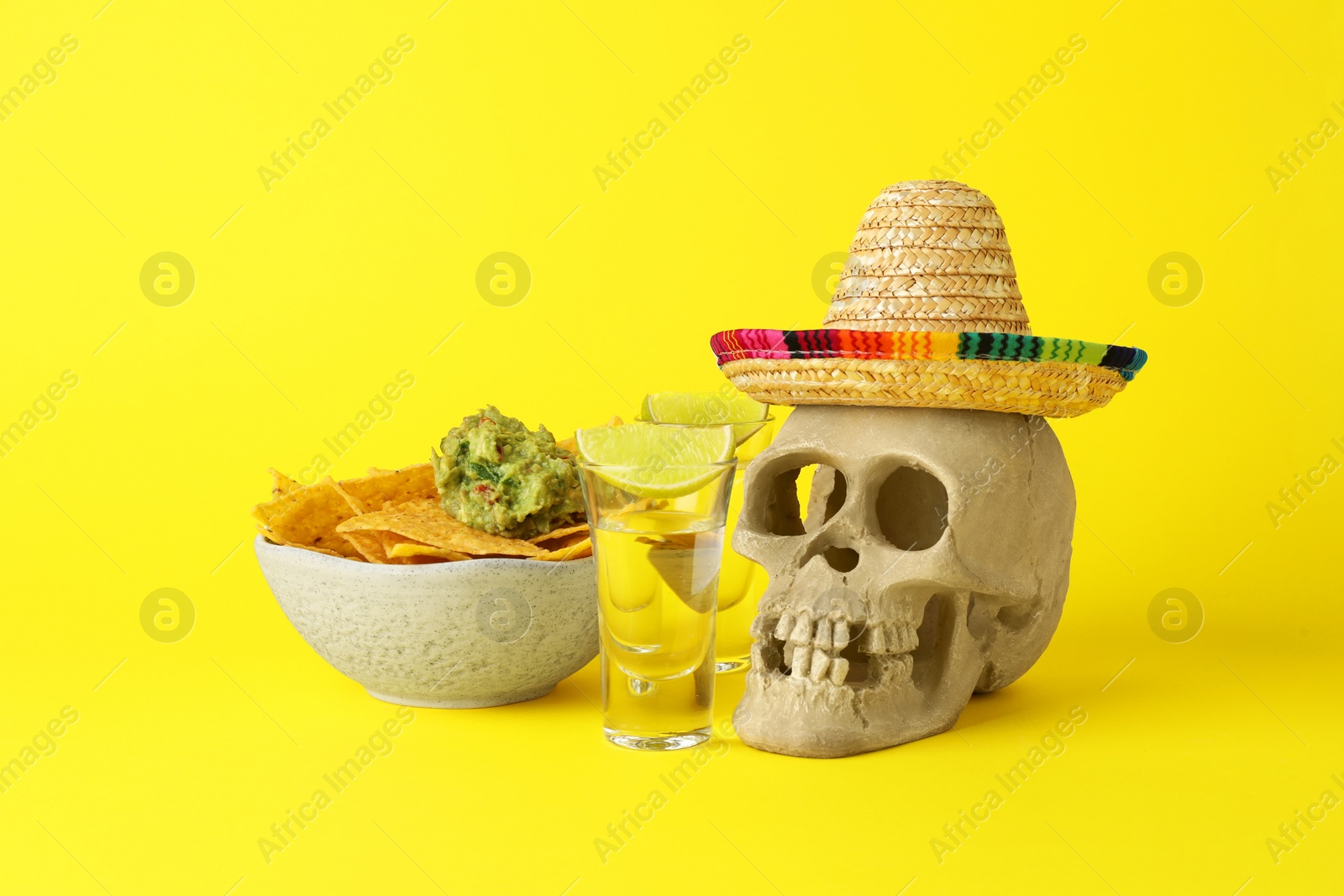 Photo of Mexican sombrero hat, human scull, tequila, nachos chips and guacamole in bowl on yellow background