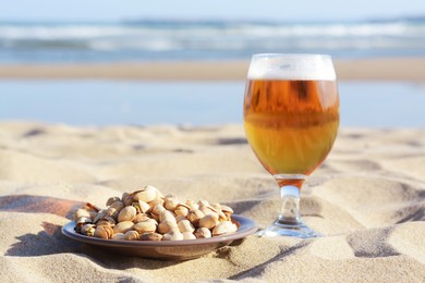 Glass of cold beer and pistachios on sandy beach near sea, space for text