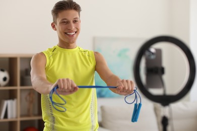 Photo of Smiling sports blogger holding skipping rope while streaming online fitness lesson with smartphone at home