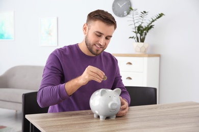 Photo of Young man putting coin into piggy bank at table indoors