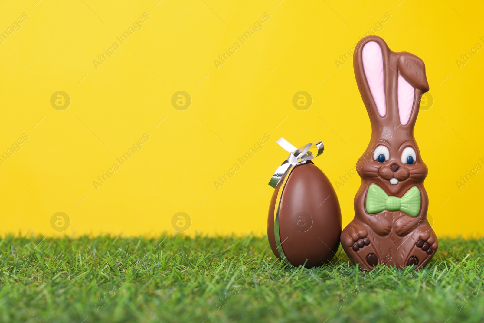 Photo of Easter celebration. Cute chocolate bunny and egg on grass against yellow background, space for text