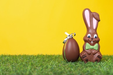 Photo of Easter celebration. Cute chocolate bunny and egg on grass against yellow background, space for text