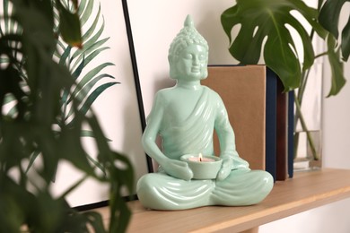 Photo of Ceramic Buddha sculpture with burning candle and books on wooden shelf indoors. Interior decor