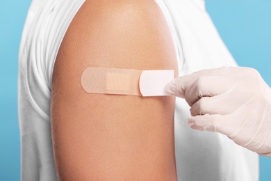 Photo of Doctor sticking plaster on man's arm after vaccination against light blue background, closeup
