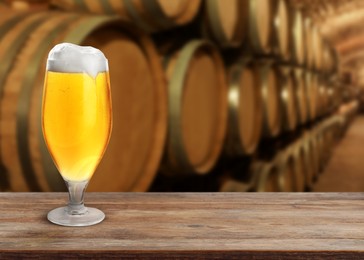 Glass of tasty beer on wooden table in cellar with large barrels, space for text