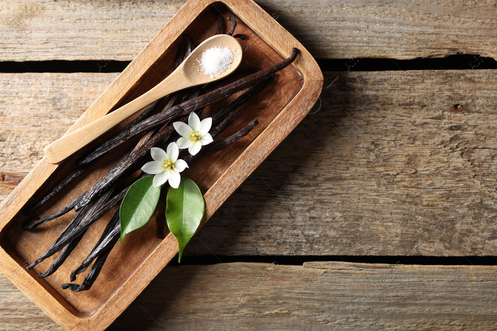 Photo of Vanilla pods, flowers, leaves and spoon with sugar on wooden table, top view. Space for text