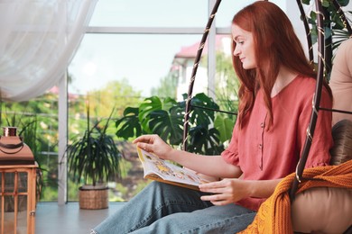 Photo of Happy young woman reading magazine in egg chair at indoor terrace