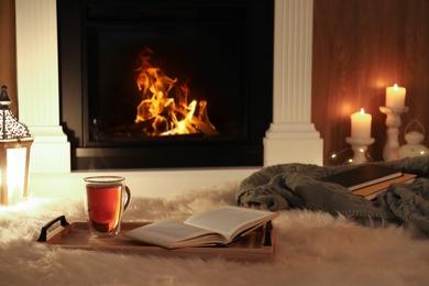 Photo of Cup of tea and book on fuzzy rug near fireplace at home. Cozy atmosphere