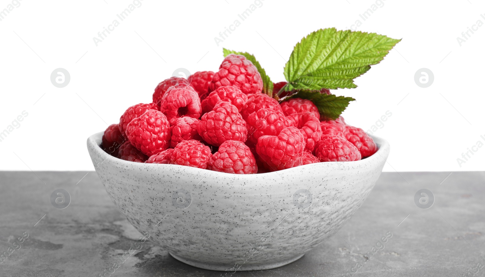 Photo of Bowl of fresh ripe raspberries with green leaves on grey table against white background