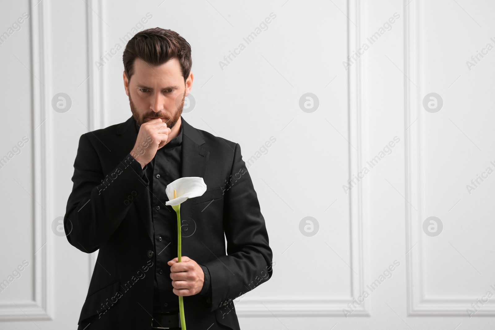 Photo of Sad man with calla lily flower near white wall, space for text. Funeral ceremony