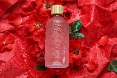 Photo of Wet bottle of essential oil and flowers on red crumpled paper, flat lay