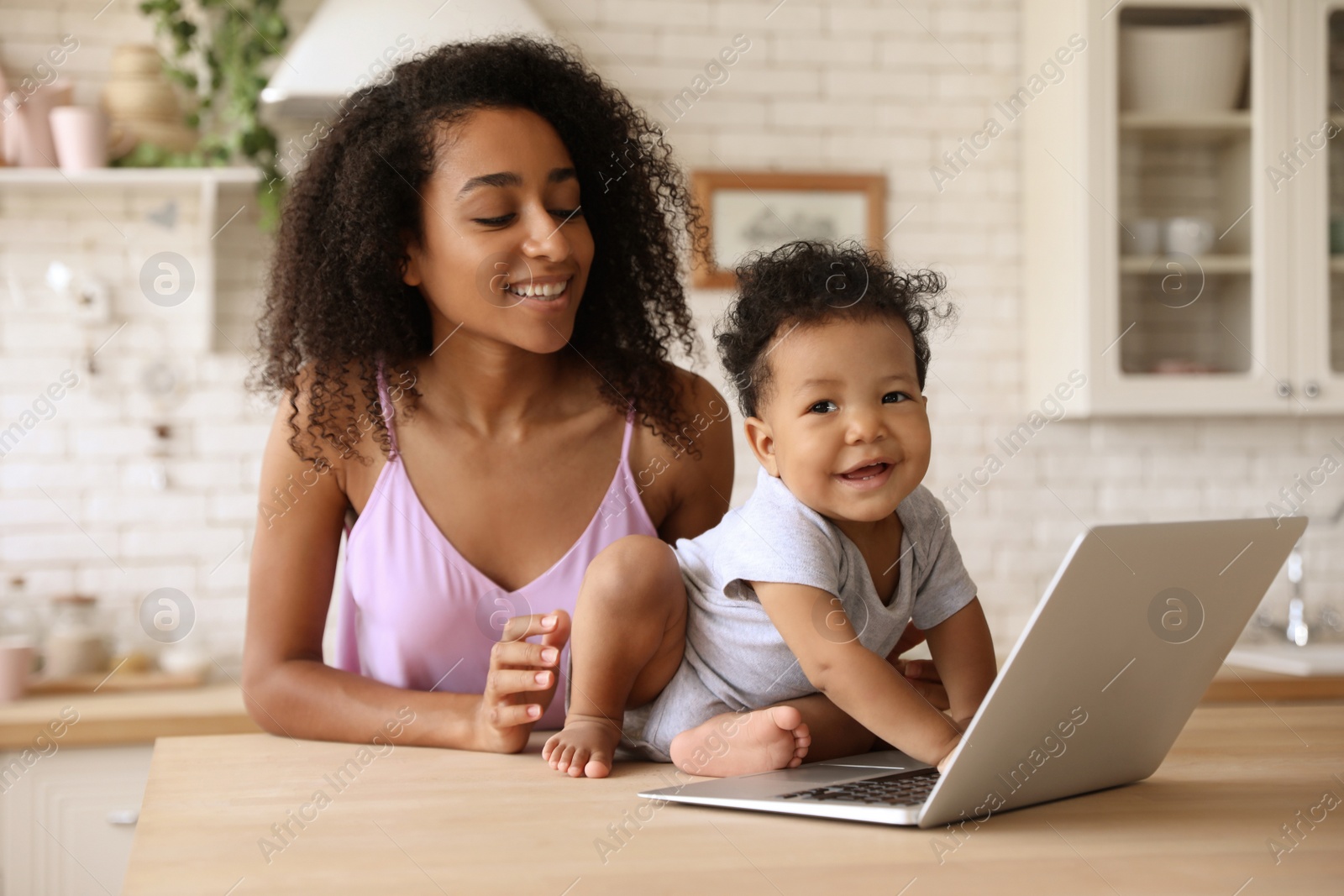 Photo of African-American woman and her baby with laptop in kitchen. Happiness of motherhood