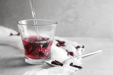 Making aromatic hibiscus tea. Pouring water into glass with dried roselle calyces and spoon on light table, space for text