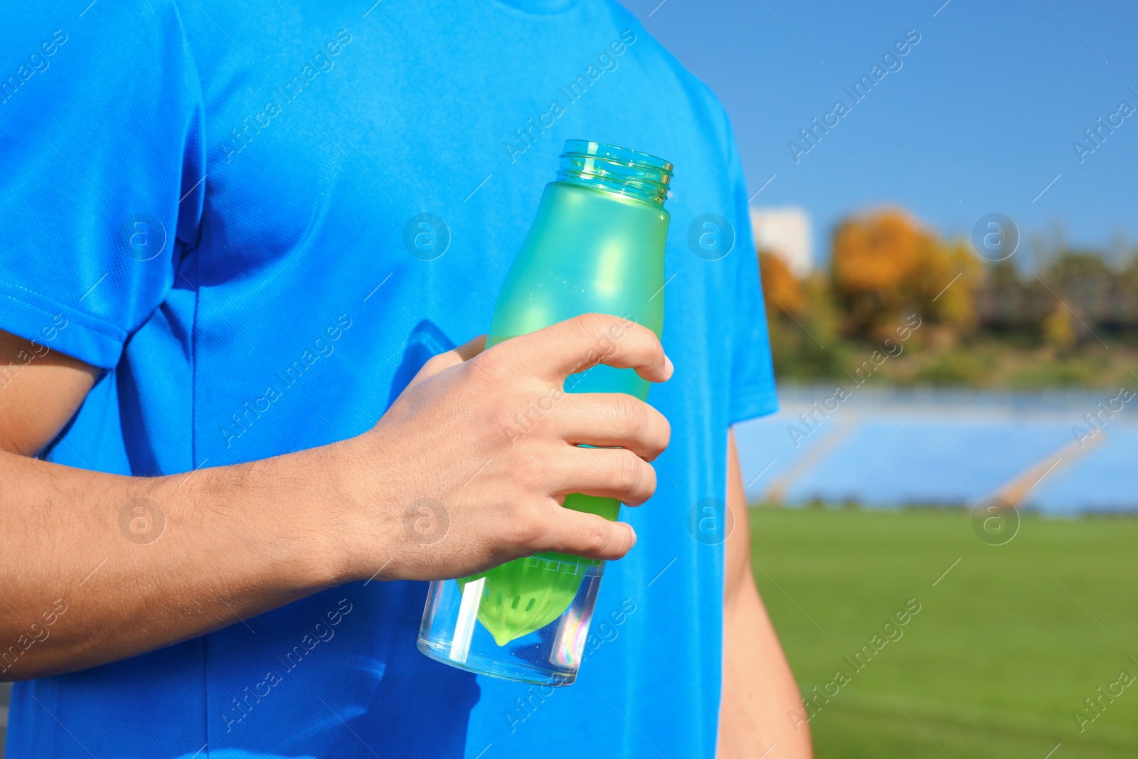 Photo of Sporty man holding bottle of water at stadium on sunny day, closeup