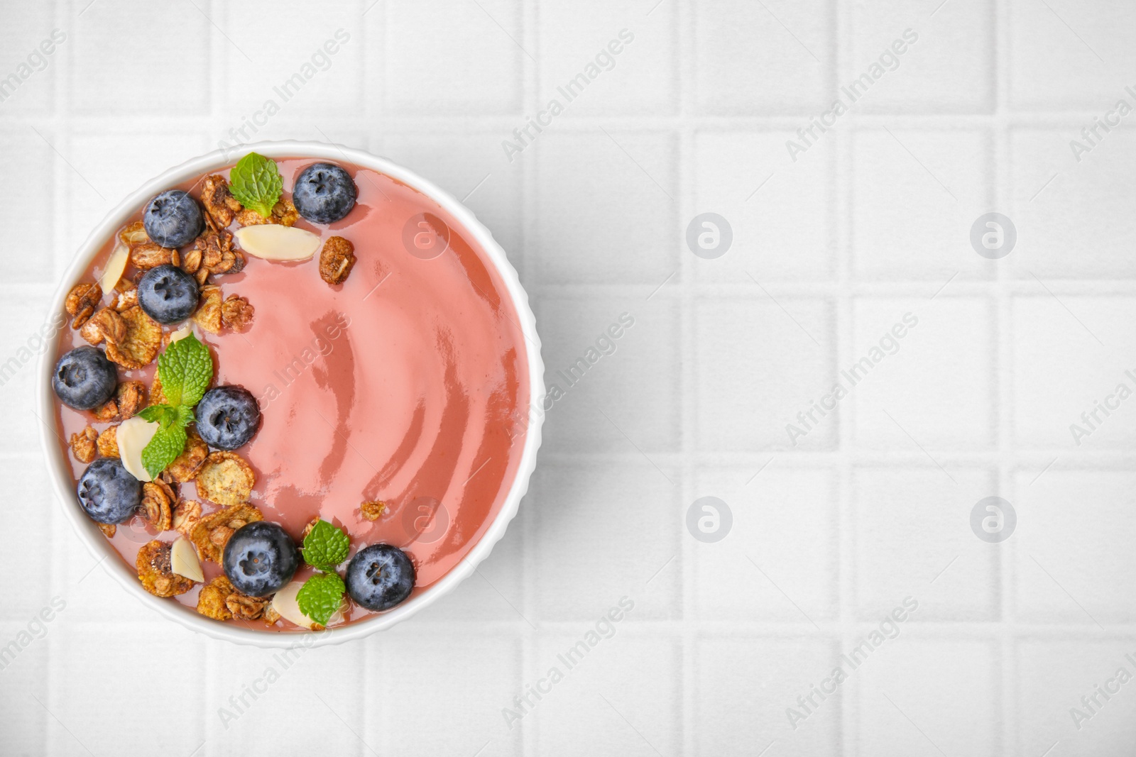 Photo of Bowl of delicious smoothie with fresh blueberries and granola on white tiled table, top view. Space for text