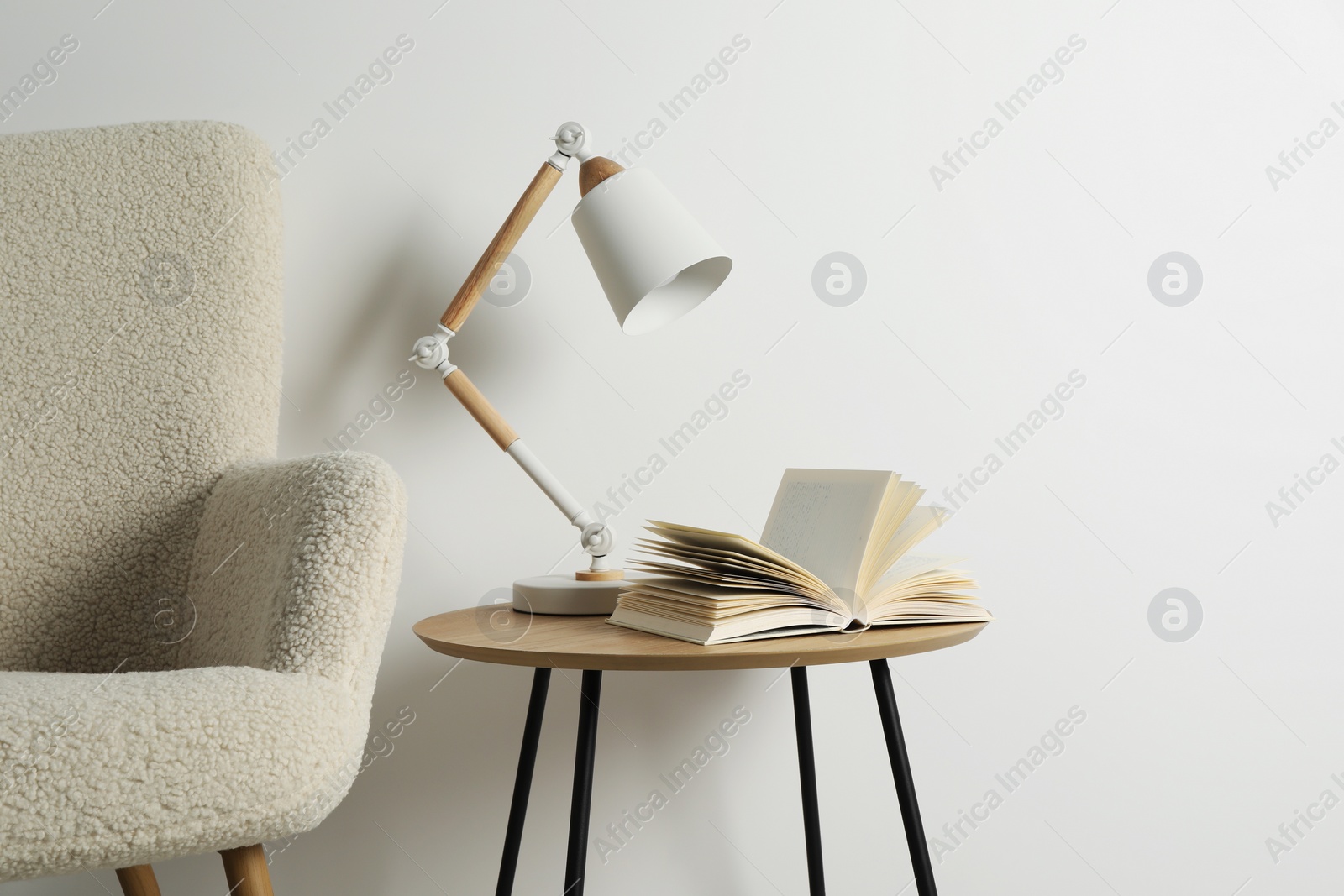 Photo of Stylish lamp with open book on wooden coffee table and soft armchair near white wall. Interior design