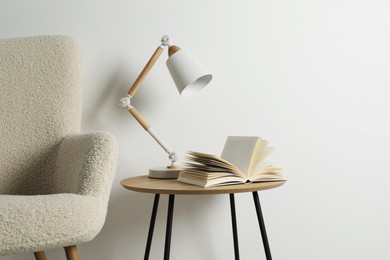 Photo of Stylish lamp with open book on wooden coffee table and soft armchair near white wall. Interior design