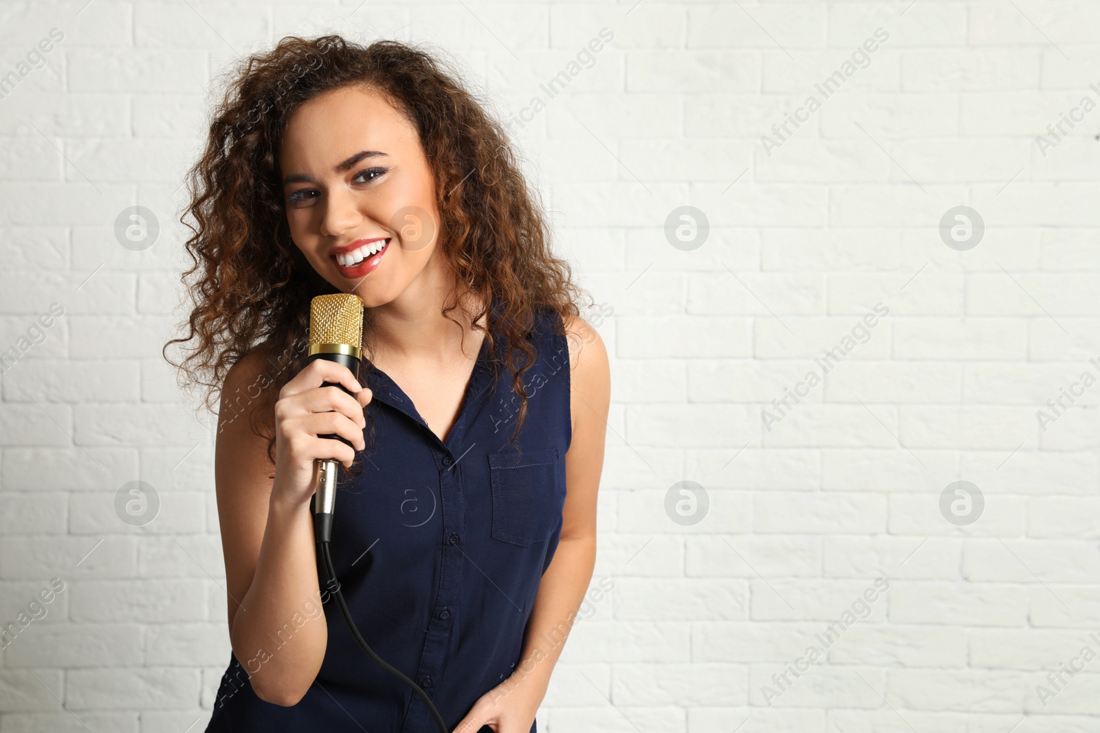 Photo of Portrait of curly African-American woman in blouse posing with microphone near brick wall. Space for text