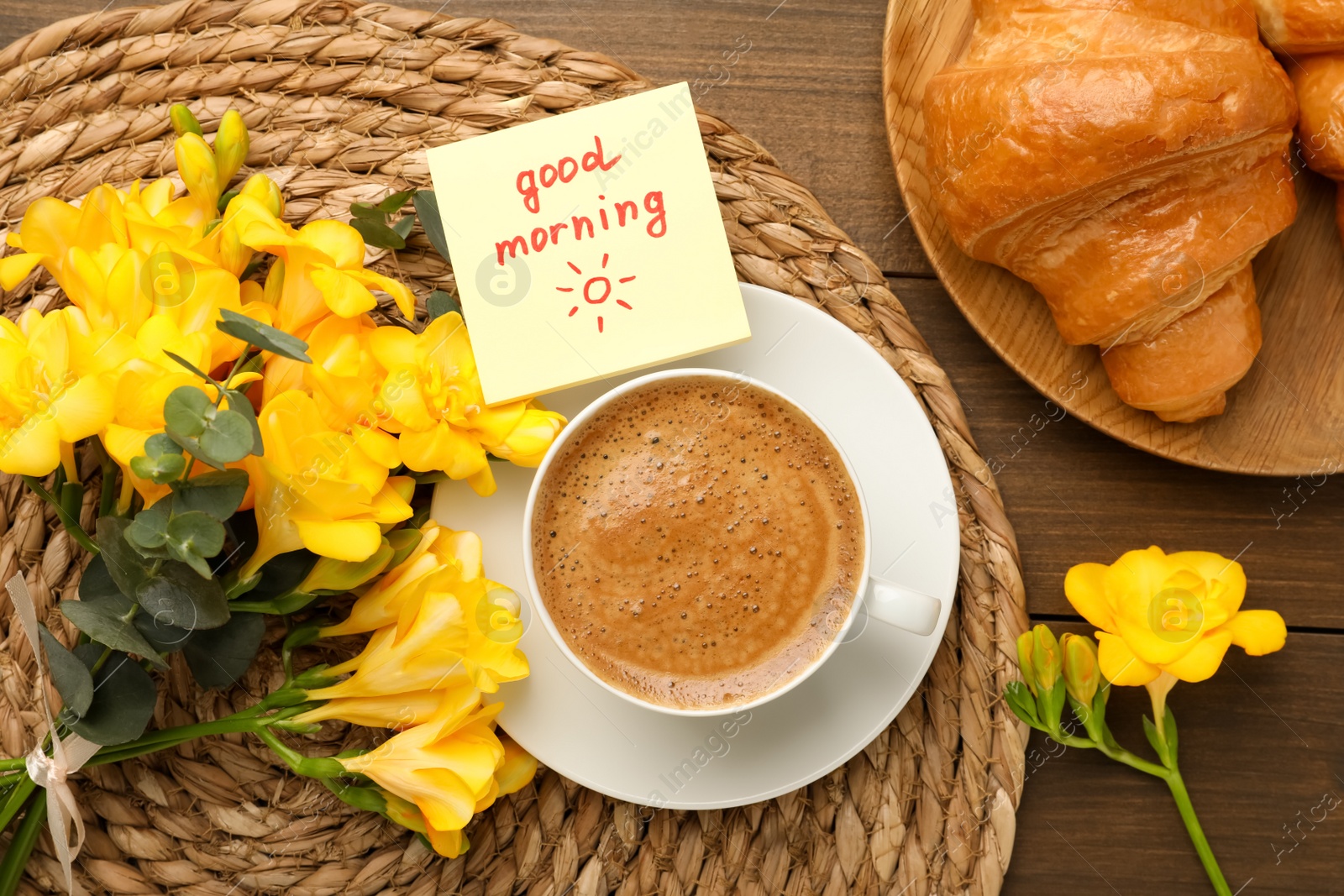 Photo of Cup of aromatic coffee with croissant, beautiful yellow freesias and Good Morning note on wooden table, flat lay