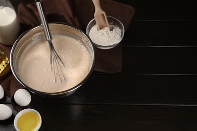 Photo of Dough, metal whisk in bowl and ingredients on dark wooden table, flat lay. Space for text