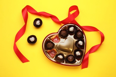 Photo of Heart shaped box with delicious chocolate candies and ribbon on yellow background, flat lay