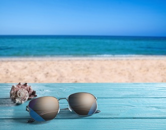 Shell and stylish sunglasses on turquoise wooden table near sea with sandy beach, space for text