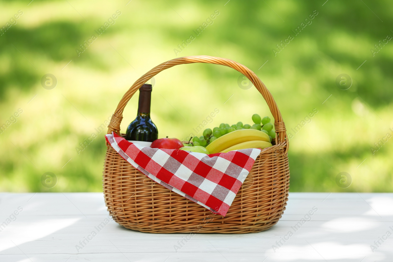 Photo of Wicker basket with blanket, wine and food on table in park. Summer picnic