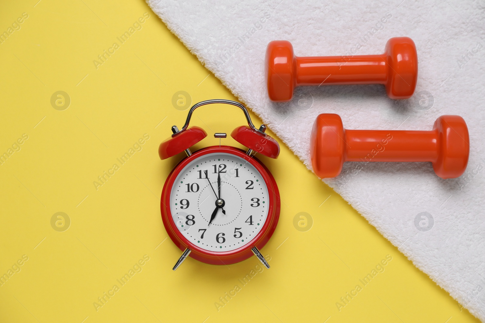 Photo of Alarm clock, towel and dumbbells on yellow background, flat lay. Morning exercise