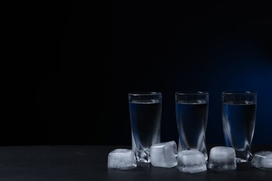 Photo of Shot glasses of vodka with ice cubes on black table against dark background. Space for text
