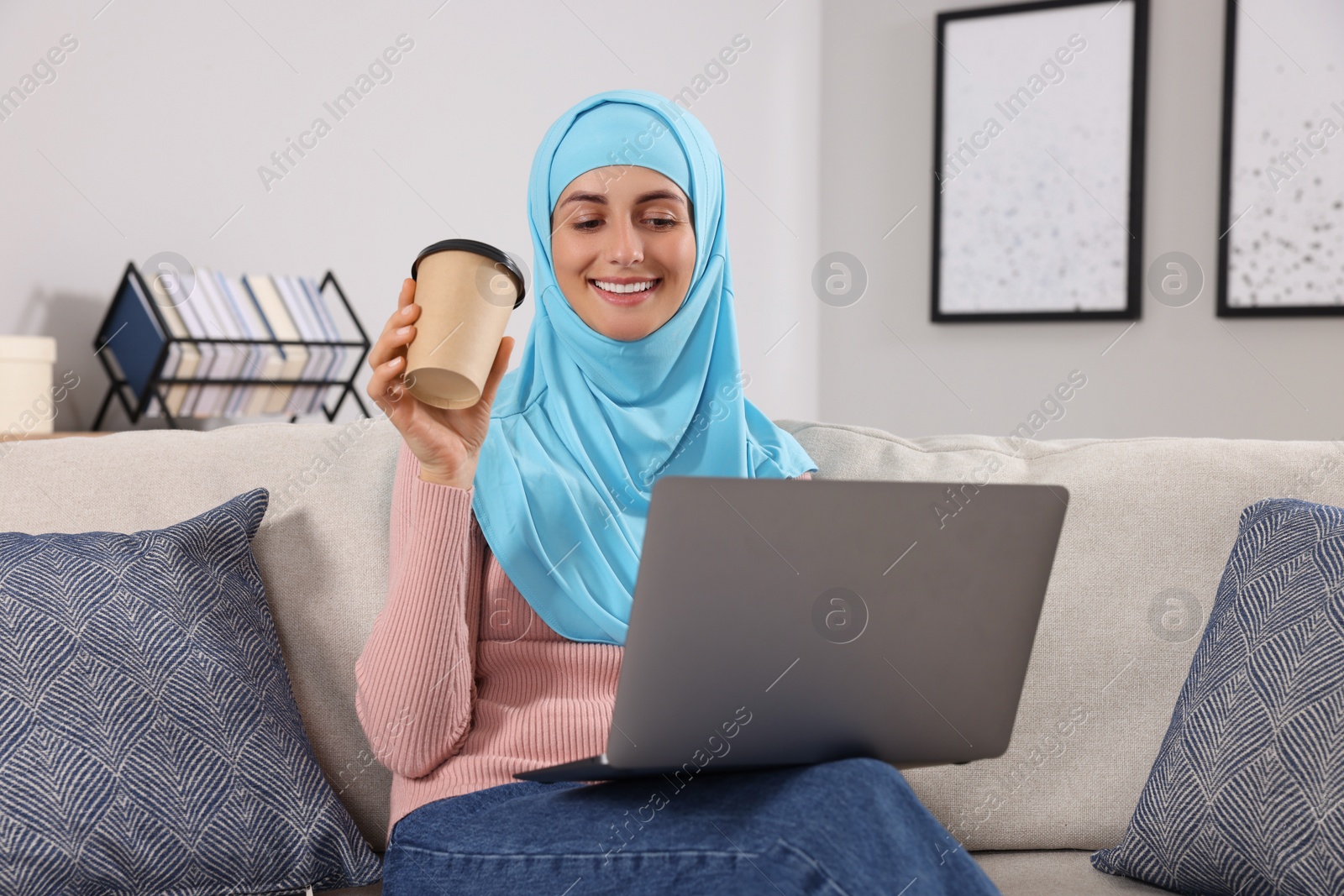 Photo of Muslim woman with cup of coffee using laptop at couch in room