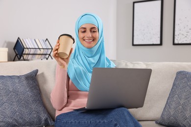 Muslim woman with cup of coffee using laptop at couch in room