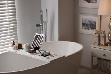 Photo of White wooden tray with tablet, spa products and burning candles on bathtub in bathroom