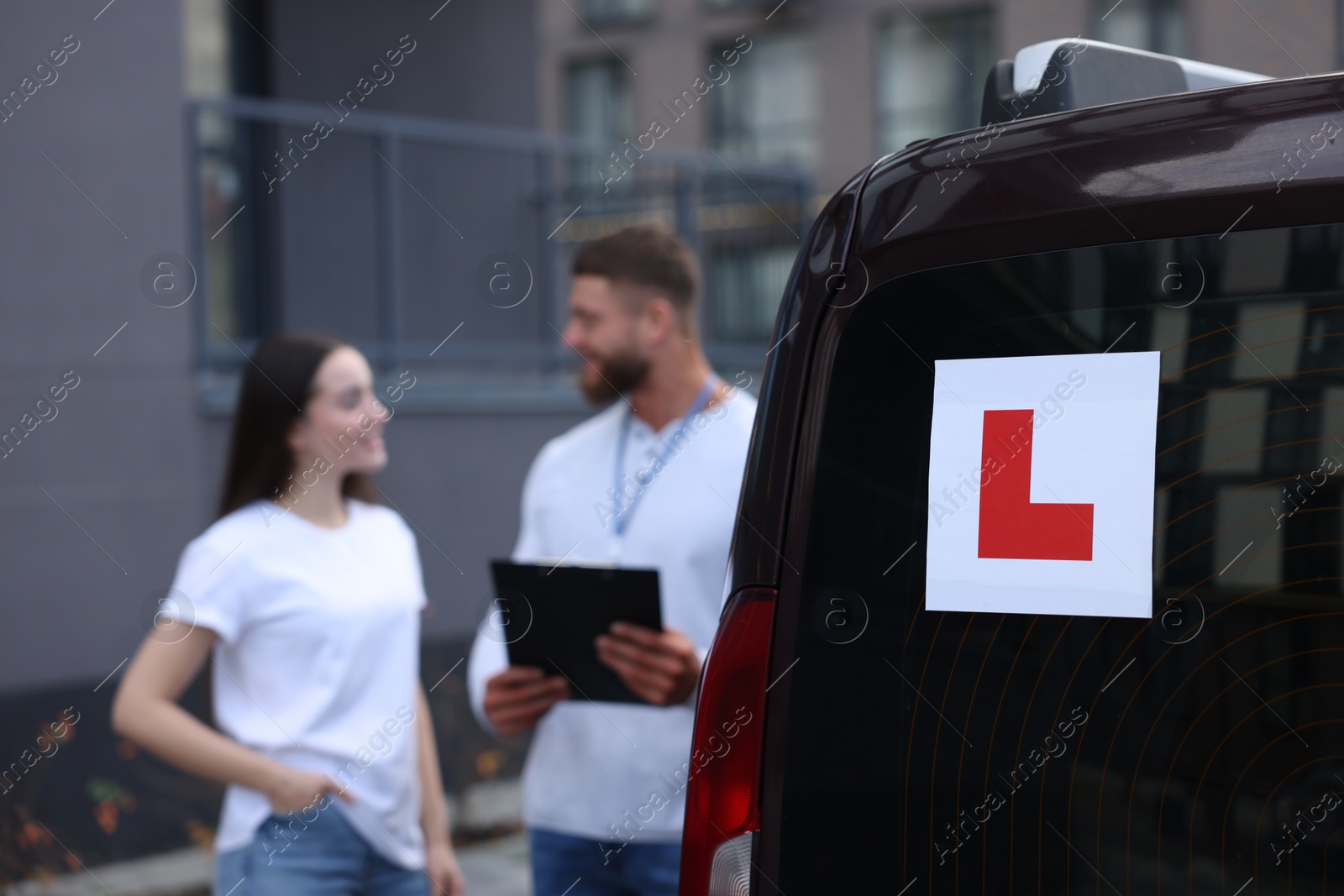 Photo of Learner driver and instructor with clipboard near car outdoors, selective focus on L-plate. Driving school