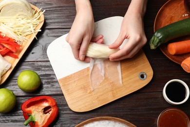 Photo of Making delicious spring rolls. Woman wrapping fresh vegetables into rice paper at wooden table, flat lay