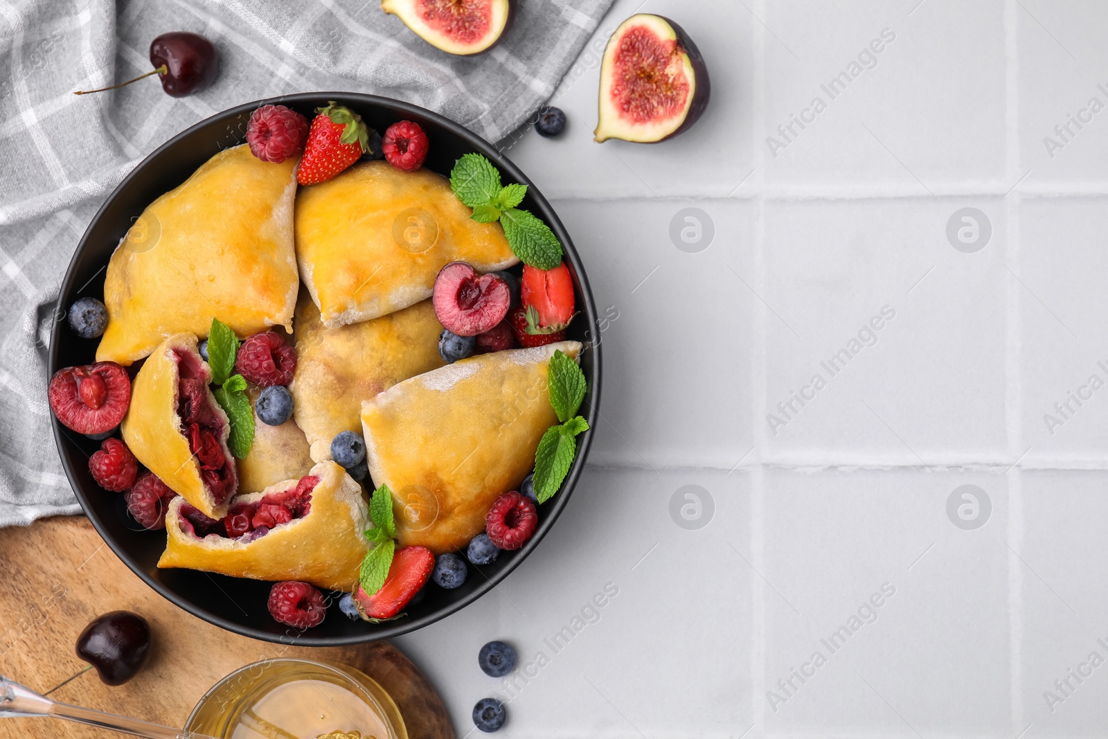 Photo of Bowl with delicious samosas, berries, fig and mint leaves on white tiled table, flat lay. Space for text