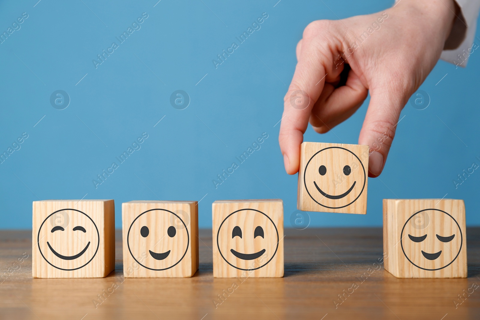 Image of Woman arranging cubes with different emoticons in row on wooden table against light blue background, closeup