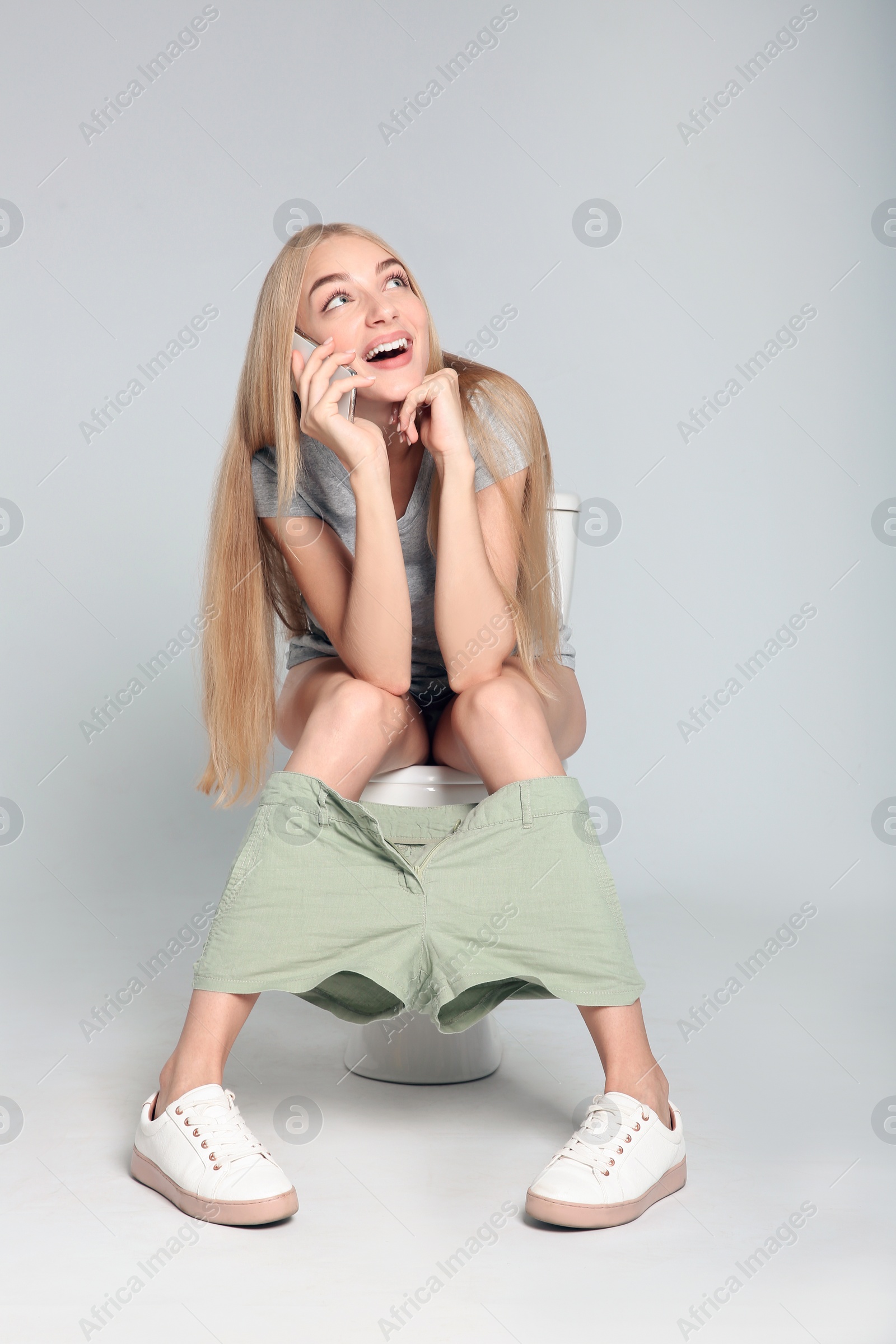 Photo of Young woman with mobile phone sitting on toilet bowl against gray background