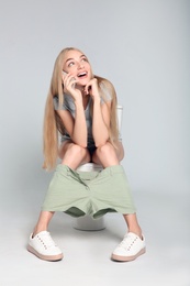 Young woman with mobile phone sitting on toilet bowl against gray background