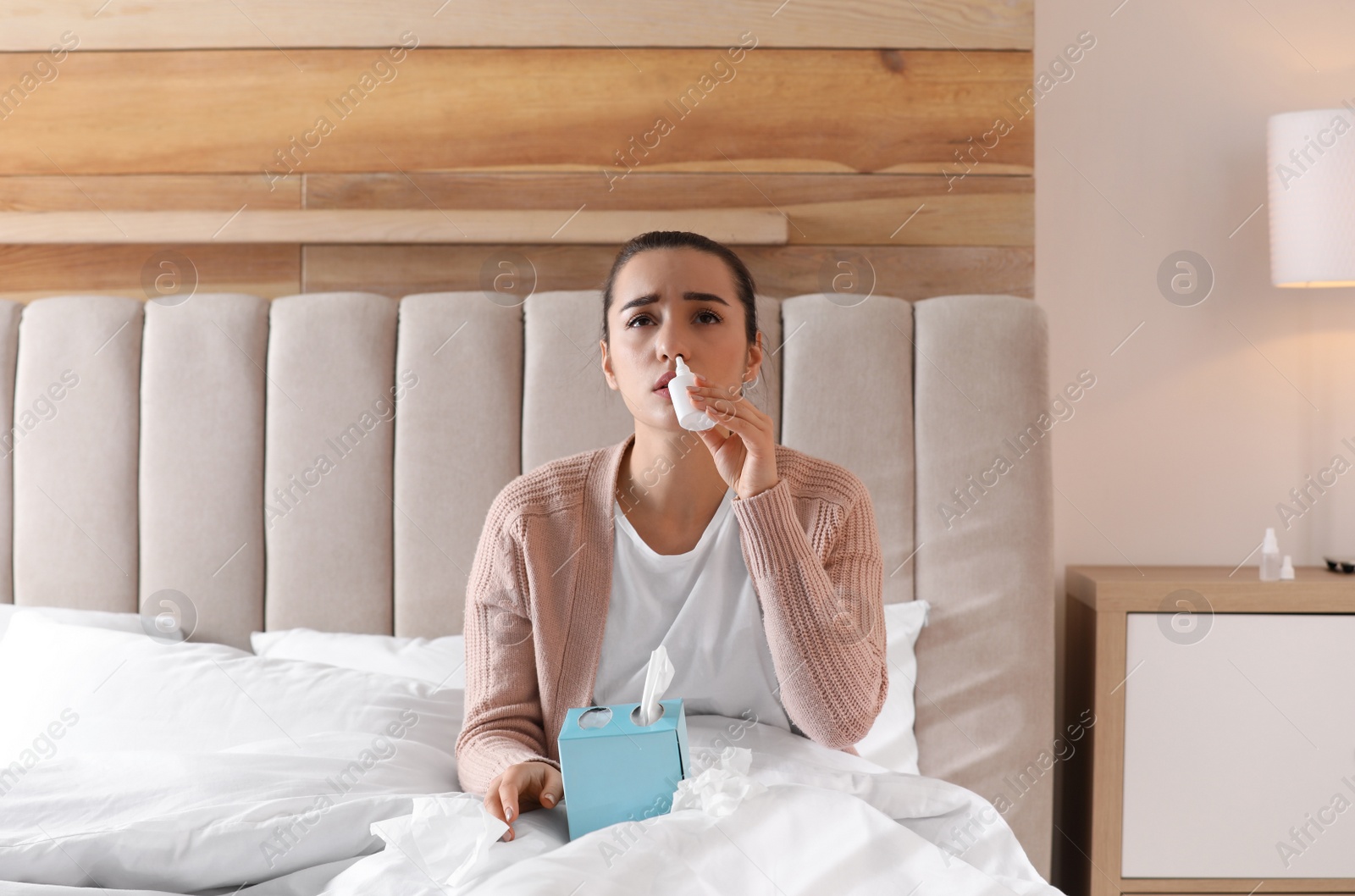 Photo of Sick young woman with napkins and nasal spray in bed at home