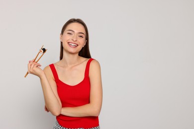 Photo of Happy young woman holding sushi roll with chopsticks on light background. Space for text
