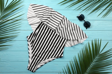 Stylish striped swimsuit, sunglasses and tropical palm leaves on light blue wooden background