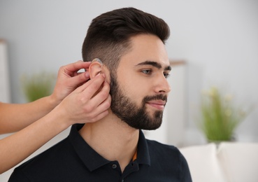Photo of Young woman putting hearing aid in man's ear indoors