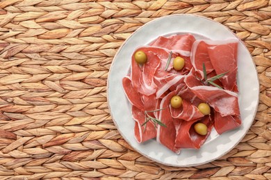 Photo of Slices of tasty cured ham, olives and rosemary on wicker mat, flat lay. Space for text