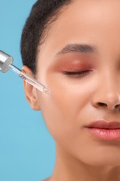 Woman applying serum onto her face on light blue background, closeup