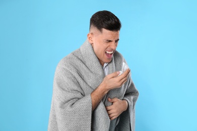 Photo of Man with blanket sneezing on light blue background. Runny nose