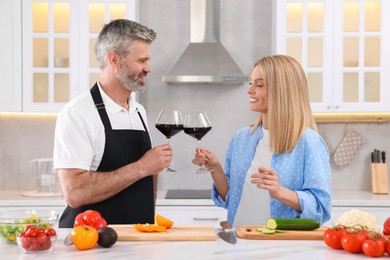 Photo of Happy affectionate couple with glasses of wine cooking together at white table in kitchen